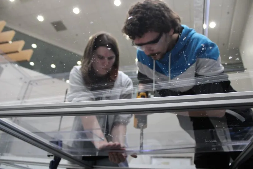 Natalie holding two sheets together while Neil drills through the polycarbonate. Natalie is wearing a grey jacket with rolled up sleeves, Neil is wearing a blue and black jacket and safety glasses.