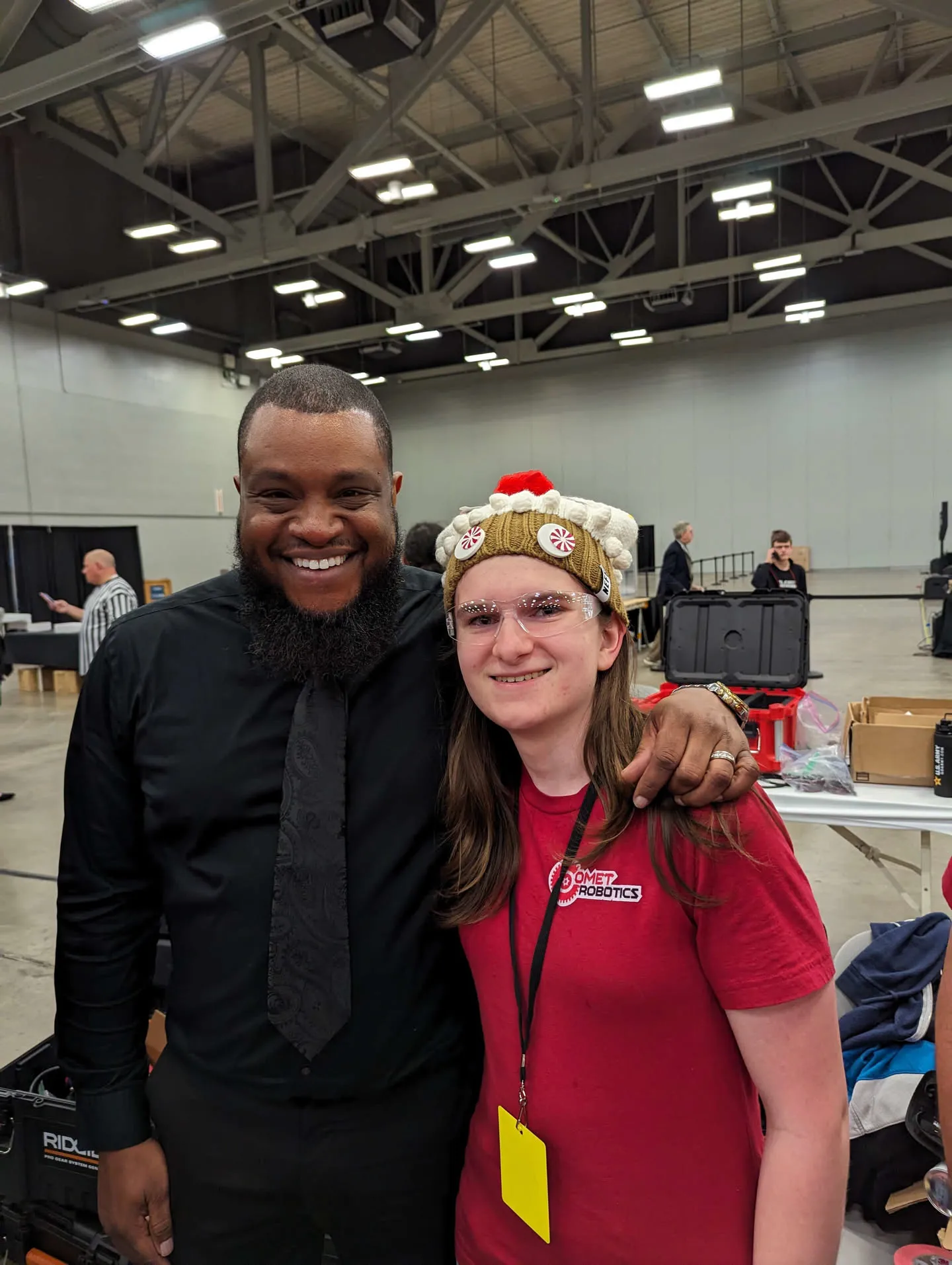 Faruq grinning in a black button up shirt and tie, with an arm over Natalie who is wearing a red shirt with the Comet Robotics logo, a cupcake beanie and safety glasses